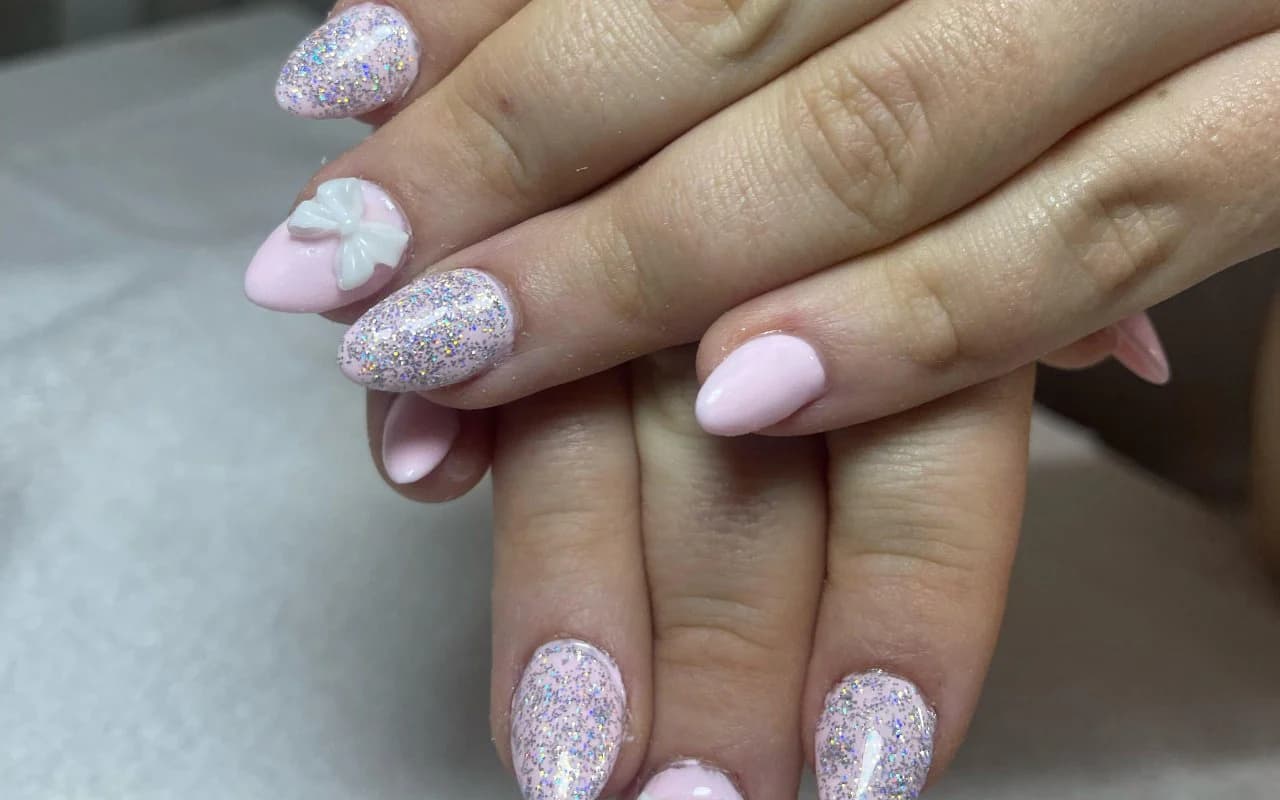 Baby pink almond gel nails with iridescent glitter and a sculpted 3D bow