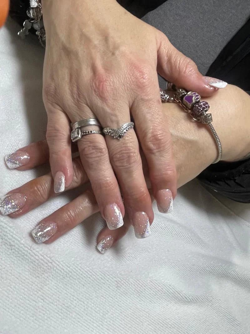 Natural nude nails with silver glitter tips, worn with stacked rings and a charm bracelet